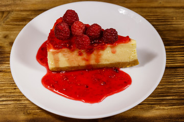 Piece of tasty New York cheesecake with raspberries and raspberry jam in a white plate on wooden table
