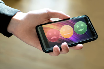 Hand holding smartphone with rating icons on touchscreen happy neutral and sad faces for customer satisfaction service quality online evaluation and survey. Modern interface feedback concept.