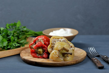 Bulgarian peppers stuffed with rice and meat on a wooden plate. Cutlery next to the plate. In the background, parsley, a bowl with sauce. Gray background. Close-up.
