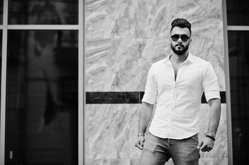 Stylish tall arabian man model in white shirt, jeans and sunglasses posed at street of city. Beard attractive arab guy.
