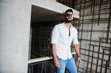 Stylish tall arabian man model in white shirt, jeans and sunglasses posed against steel wall indoor. Beard attractive arab guy.