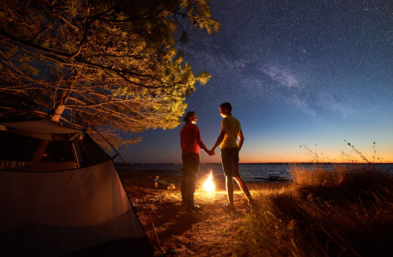 Night camping at lake under blue starry sky. Back view of young tourists, man and woman standing, holding hands at campfire on clear water and city lights on distant shore background. Tourism concept