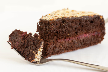 One slice of chocolate cake filled with jam on white background. One bite is on fork.