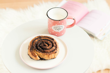 Chocolate roll on white plate, a glass of milk in a pink mug with 'Homemade with love' sign and an opened book on a white plush carpet.