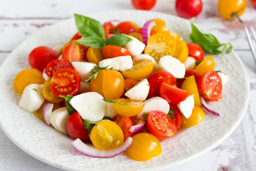 Colorful, fresh tomato salad with red and yellow cherry tomatoes, mozzarella and basil on white plate.