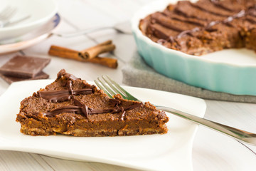One slice of chocolate pie with cinnamon served on white plate, whole cake is in the background in blue pie baking form.