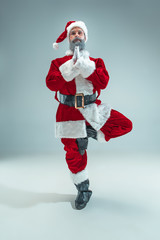 Funny serious guy with christmas hat sitting at yoga pose at studio. New Year Holiday. Christmas, x-mas, winter, gifts concept. Man wearing Santa Claus costume on gray. Copy space. Winter sales.