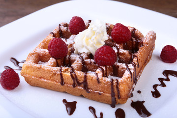 Closeup of homemade Belgian waffles with whipped cream, chocolate sauce and raspberries on white plate.