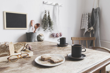 Vintage interior design of kitchen space with wooden table against white wall with simple chairs, cups of coffee and breakfast. Minimalistic concept of kitchen space.