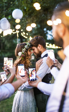 Guests with smartphones taking photo of bride and groom at wedding reception outside.