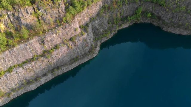 (Drohne) Schwenk &uuml;ber Felswand eines Steinbruchs mit blauem See im Vordergrund