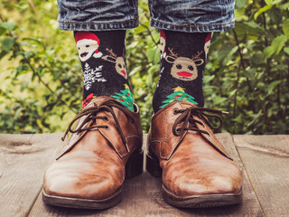 Men's legs in stylish shoes, bright, variegated socks with Christmas and New Year's patterns on the wooden terrace on the background of green trees. Beauty, fashion, elegance