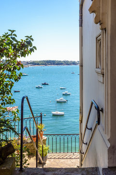 The doorstep of a villa in Dinard in Brittany, France, with a balcony down the stairs overlooking the estuary of the river Rance with boats moored in the clear waters in the distance.