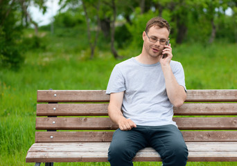 The caucasian cheerful man, 27 years old, with glasses and short hairstyle is talking on the mobile phone in a park. European male model is sitting on a wood bench in outdoors.