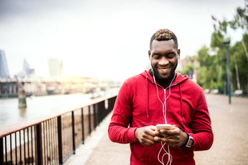 Young sporty black man runner on the bridge outside in a city, using smartphone.