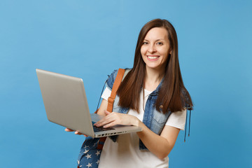 Young attractive happy woman student in denim clothes with backpack holding and using working on laptop pc computer isolated on blue background. Education in college. Copy space for advertisement.