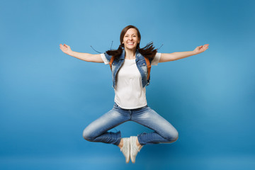 Full length portrait of young smiling woman student in denim clothes jumping spreading hands putting feets together isolated on blue background. Education in university. Copy space for advertisement.