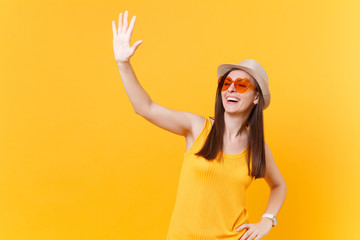 Portrait of smiling young woman in straw summer hat, orange glasses waving and greeting hand as notices someone, copy space isolated on yellow background. People lifestyle concept. Advertising area.