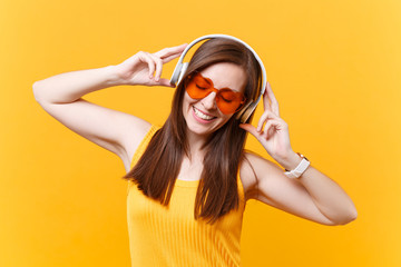 Portrait of calm smiling young woman in orange glasses listening music in headphones copy space isolated on yellow background. People sincere emotions, lifestyle concept. Advertising area. Rest, relax