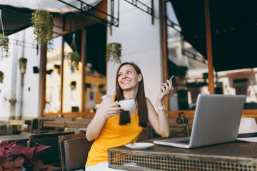 Fototapeta premium Happy girl in outdoors street coffee shop cafe sitting at table with laptop pc computer, texting message on mobile phone, drink cup tea in restaurant during free time. Mobile office freelance concept.