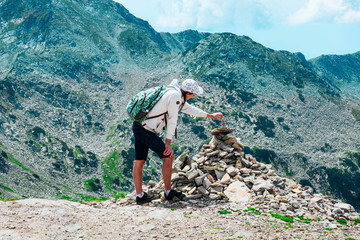 A young man hiker, reached the mountain top, building pyramid of rocks, beautiful alpine landscape, summertime.