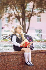 A small cheerful blue-eyed girl, a first-class woman with winded hair with bows and glasses in school uniform with a briefcase and book on September 1, is sitting in the school park near the fountain