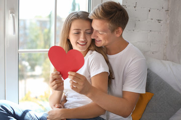 Happy young couple with red heart sitting near window at home
