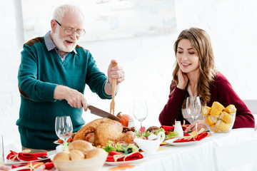happy senior man slicing delicious turkey for thanksgiving celebration while his adult daughter sitting near at table