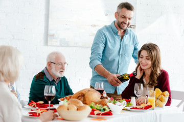 happy adult man pouring wine into glass at served table while family having thanksgiving celebration