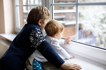 Happy adorable kid boy and cute baby girl sitting near window and looking outside on snow on...