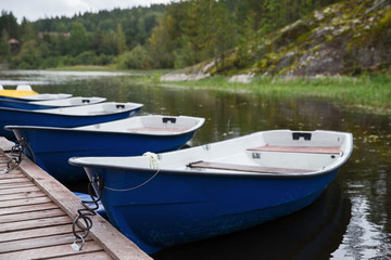 Blue empty boats on the lake along the wooden pier closeup, cloudy autumn sky and forest on background .  Recreational row boats. Boat rental