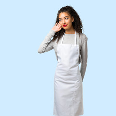 Young girl with apron listening to something by putting hand on the ear on isolated blue background