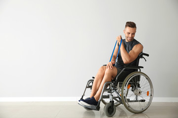 Fototapeta na wymiar Sporty man training with elastic band while sitting in wheelchair against light wall