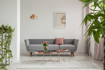 Close-up of leaves in an elegant living room interior with a grey couch and copper tables decorated with roses. Real photo - obrazy, fototapety, plakaty
