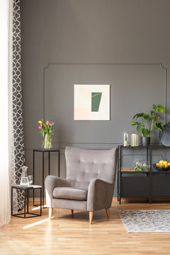 Flowers on table behind beige armchair in grey living room interior with poster. Real photo