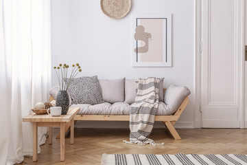 Wooden table next to beige settee with blanket in bright living room interior with poster. Real photo