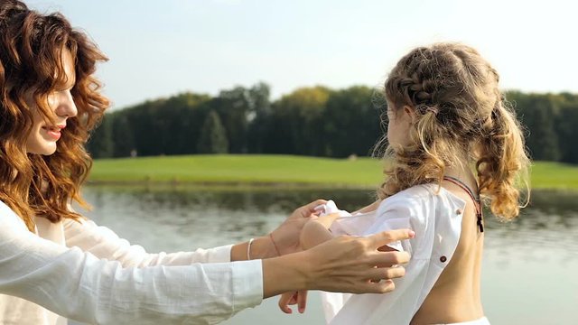 Happy moyher and daugher in the park near the pond. Mother undressing little cute daughter and prepearing for swimming in the lake. Steadycam shot.