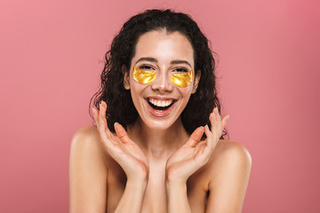 Beauty portrait of european shirtless woman with long brunette hair wearing cosmetic patches under her eyes, isolated over pink background