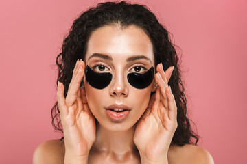 Portrait of beautiful nude woman with long brunette hair wearing cosmetic patches under her eyes, isolated over pink background