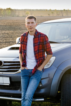 A young man in a red shirt is standing near the road next to his car