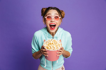 Photo of european girl 20s in sunglasses laughing and holding bucket with popcorn, isolated over violet background in studio - obrazy, fototapety, plakaty