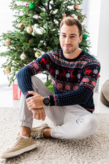 handsome man in winter sweater sitting near christmas tree
