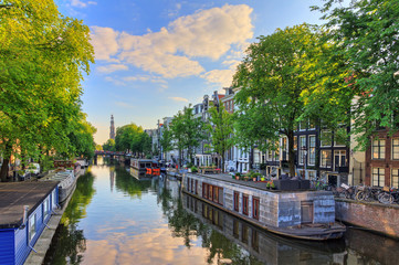 Obraz premium Łodzie mieszkalne nad kanałem Prinsengracht światowego dziedzictwa UNESCO z Westerkerk (kościół zachodni) w letni poranek z błękitnym niebem i chmurami oraz lustrzanym odbiciem w Amsterdamie, Holandia