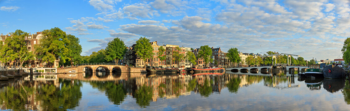 Beautiful panoramic panorama of the river Amstel and the skinny bridge (Magere brug) in Amsterdam, the Netherlands, on a sunny summer morning with some clouds and a mirror reflection