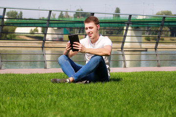 Naklejka na ściany i meble Handsome boy using tablet and headphones, drinking coffee to go, man sitting on grass and enjoying nice sunny day by river side. Model photo-shoot, urban fashion, lifestyle. White T-shirt and jeans.