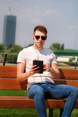 Obraz premium Handsome boy wearing sunglasses using tablet and headphones, sitting on a bench and drinking coffee to go. Handsome student immersed in social media. Background scenery bridge, river and green grass.