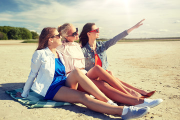 Fototapeta premium summer vacation, holidays, travel and people concept - group of smiling young women in sunglasses sitting on beach blanket and pointing finger to something