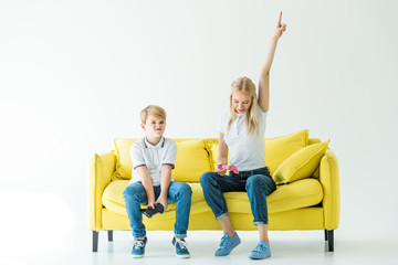 mother showing one hand up after winning video game, son sitting upset on yellow sofa on white