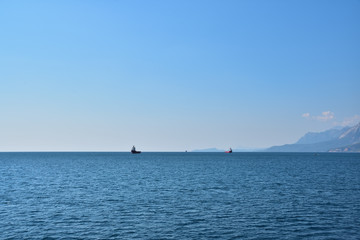Two ships for transporting containers at anchorage in the sea, against the backdrop of the coastline, mountains, blue sky