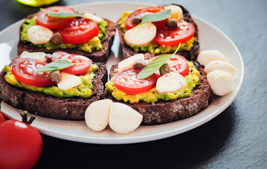 Sandwich toasts with tomatoes cherry, mozzarella, avocado, basil and olive oil. Side view on a dark stone dish.